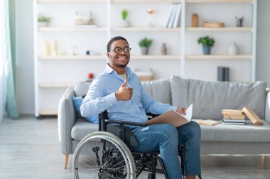 Tekerlekli sandalyedeki sakat, elinde not defteri olan mutlu siyahi adam parmağını kaldırıyor ve evde gülümsüyor. Milenyum Afrikalı Amerikalı engelli adam içeride mi çalışıyor yoksa çalışıyor mu?