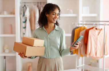 Sırıtkan girişimci arka planında elbise askısı olan modern moda stüdyosunda akıllı telefonunu kontrol ederken karton kutular taşıyordu.