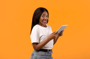 Modern dijital tabletli, turuncu stüdyo arka planında poz veren pozitif, genç siyahi kadın, kopyalama alanı. Neşeli Afrikalı kadın cep telefonu uygulamasını ped, teknoloji ve eğlence konseptinde kullanıyor.