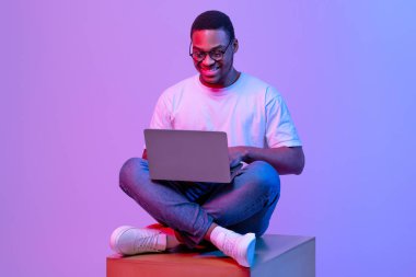 Yakışıklı Siyah Programcı Laptop Bilgisayar kullanıyor, Neon Işıklı Büyük Küp 'te oturuyor, Gözlüklü Neşeli Afrikalı Amerikalı Adam Mor Stüdyo Arkaplanı Üzerinde Çalışıyor veya Çalışıyor, Boşluğu Kopyala