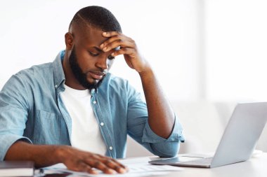 İş sorunları işte. Hüsrana uğramış Afrikalı Amerikalı işadamı ofiste evrak okuyor, masasında oturuyor ve mali raporları kontrol ediyor, yorgun ve yorgun hisseden siyahi bir adam,