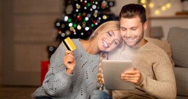 Tatil teklifleri. Genç ve mutlu alışveriş düşkünleri dijital tablet, ev içi, fotokopi alanı üzerinden alışveriş yapıyorlar. Kredi kartı ve bloknotu olan mutlu çift internetten alışveriş yapıyor, hediyeler alıyor.