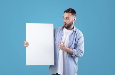 Mavi stüdyo arka planındaki beyaz boş posteri gösteren yeni nesil bir adam. Reklam tasarımın için bir model. Yakışıklı beyaz adam, boş afişi kopyalama alanıyla gösteriyor