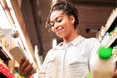 Afro-Amerikalı bir kadın market alışverişi yapıyor yiyecek alıyor, indirimler yapıyor ve markette alışveriş arabasıyla kuponlar duruyor. Kadın müşteriler marketten alışveriş yapar. Alıcı uygulaması