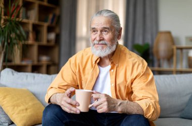 Happy grey-haired bearded senior man in stylish casual outfit drinking coffee at home, holding cup and looking at copy space for advertisement or text, living room interior