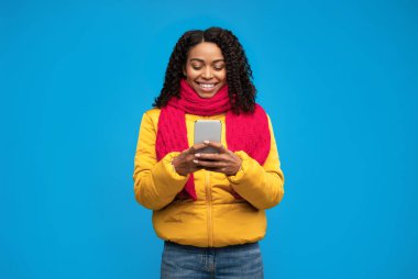 Neşeli Afro-Amerikalı Kız İnternet 'te Mesaj Kullanıyor Kış Giysileri Giyiyor Mavi Arkaplanda Stüdyoda. Mobil Uygulama Kavramı