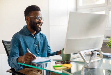 Positive black businessman writing notes in notebook while watching webinar video course, looking at computer monitor and smiling, sitting at workplace in office