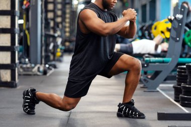 Modern spor salonunda çömelme hareketleri yapan siyahi bir sporcu, Afrika asıllı Amerikan kaslı genç vücut geliştirici, bacak kasları üzerinde çalışıyor, fotokopi alanı olan panorama, yan görüş.
