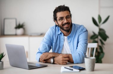 Serbest meslek hayatı. Gayri resmi giyinen yakışıklı Hintli adam, ofisteki laptop masasında otururken kameraya gülümsüyor, neşeli Batılı genç adam bilgisayarla uzaktan çalışmaktan hoşlanıyor, Boşluğu Kopyala