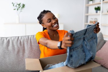 İnternetten kıyafet sipariş etmek. Gülümseyen siyah kadın müşteri paket kutusunu açıyor, evdeki koltukta yeni kot pantolonlara bakıyor. Genç Afro kadın internet alışveriş ve teslimat hizmetlerinden memnun.