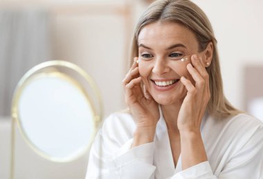 İpek bornozlu mutlu sarışın bayan nemlendirici göz kremi kullanıyor, aynaya bakıyor ve gülümsüyor, yüz bakımından keyif alıyor, yakın çekim yapıyor. Koyu halkalar, göz kırışıklıkları tedavisi