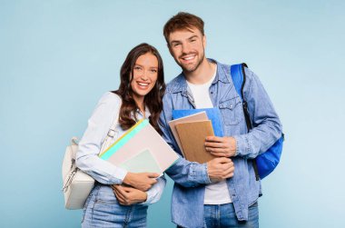 Kot pantolonlu mutlu genç çift, sırt çantalı ve kitap tutan bir adam, ve her ikisi de parlak mavi arka planda, üniversitede güne hazır klasörleri tutan bir kadın.