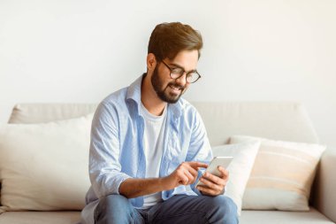 Faydalı bir uygulama. Gözlüklü yakışıklı Arap adam evde koltukta dinlenirken cep telefonu kullanıyor, Doğu Yakalı adam oturma odasında oturuyor, internete bakıyor ya da arkadaşlarıyla mesajlaşıyor, boş alan arıyor.