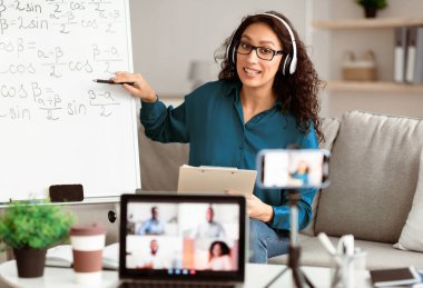 Video konferansı ve eğitim. İnternet uygulaması kullanarak çeşitli öğrencilerin uzaktan öğrendiği bir sınıf. Kulaklıklı kadın öğretmen bilgisayar ekranına bakarak ders veriyor, cep telefonuyla tripoda video kaydediyor.