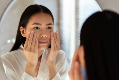 Mirror reflection of upset young asian woman in white bathrobe looking at mirror and touching her cheeks, having oily or dry skin problem, first wrinkles concept, bathroom interior, closeup photo