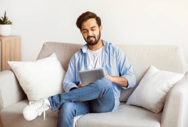 Modern Boş Zamanlar Teknolojileri. Gülümseyen Arap Adam Oturma odasında koltukta dinlenirken dijital tablet kullanıyor, Doğu Milenyum Adamı İnternet 'te alışveriş yapıyor ya da İnternet' i tarıyor, Boşluğu kopyalıyor