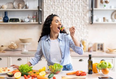 Masada organik sebzelerle salata yiyen, sağlıklı beslenmenin tadını çıkaran, açık mutfakta duran genç bir kadın. Bayan evde sebze yemeği pişirdi. Kilo verme konsepti