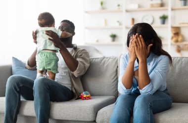 Partum bunalımı sonrası. Yorgun siyah anne yüzünü kapatıyor, evde oturmuş ağlıyor, baş ağrısı ve stres hissediyor, kocası küçük bebekle oynuyor. Ebeveynliğin zorlukları işte. Bebek mavisi