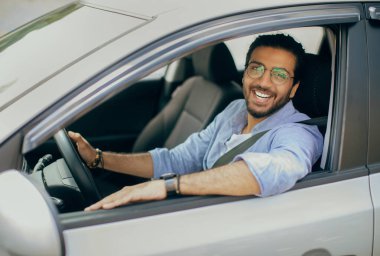 Neşeli, uzun saçlı, gözlüklü, sağ tekerli araba kullanan, açık pencereden bakan ve kameraya gülümseyen, mutlu, orta doğulu, araba sürmekten hoşlanan, uzay kopyalayan Arap adam.