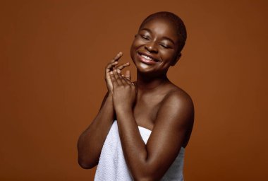 Kusursuz pürüzsüz derisi olan çekici siyah kadın doğal güzelliğinin tadını çıkartıyor, güzel Afrikalı Amerikalı kadın havluya sarılmış, kahverengi arka planda izole edilmiş, boşluğu kopyalıyor.