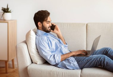 Serbest Kavram. Yakışıklı Arap adam dizüstü bilgisayar kullanıyor ve evde cep telefonuyla konuşuyor, Milenyum doğulu adam oturma odasında otururken uzaktan çalışıyor, uzak işin tadını çıkarıyor, yan görünümün tadını çıkarıyor.