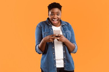 Harika bir uygulama. Neşeli Siyah Kadın Yeni Akıllı Telefon ve Mobil Uygulama Poz Veriyor Sarı Stüdyo Arkaplanının Üzerinde Duran Kameraya Gülümsüyor. Kadın Telefon Konseptinde Oyun Oynuyor