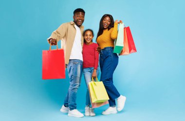 Aile alışverişi zamanı. Mutlu siyah adam, kadın ve kızları renkli alışveriş torbalarıyla birlikte poz veriyor, sarılıyor ve mavi duvardan izole edilmiş kameraya bakıyor.