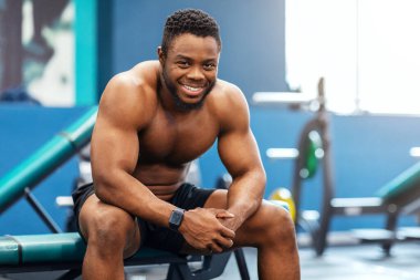 Gülümseyen Afro-Amerikan tişörtsüz vücut geliştirici modern spor salonunda antrenmandan sonra kameraya gülümsüyor. Egzersiz yaptıktan sonra dinlenen, bankta oturan, boşluğu kopyalayan mutlu siyahi adam.