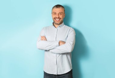 Erkek portresi. Kolları kavuşturulmuş, kameraya gülümseyen, mavi stüdyo arka planında poz veren mutlu orta yaşlı beyaz adam. Olgun erkek gülüşü.