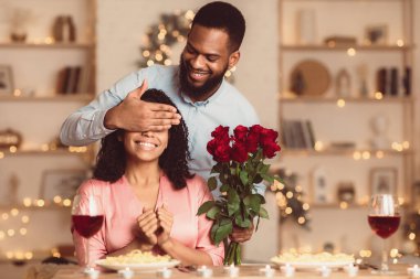 Tatille Selamlama. Gülümseyen siyah adam kadın gözlerini kapatıyor ve ona bir demet kırmızı gül veriyor, güzel bayanı şaşırtıyor. Afro-Amerikalı çift evde ya da kafede kutlama yapıyor.