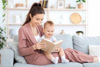 Bebekler için erken gelişim. Milenyum annesi evdeki küçük bebeğine kitap okuyor, rahat bir oturma odasında kanepede oturuyor, boş yer var.