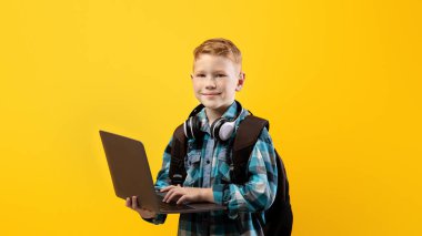 Okul eğitimindeki teknolojiler. Sırt çantası ve kulaklık takan neşeli okul çocuğu sarı stüdyo arka planında dizüstü bilgisayarı tutuyor, kopyalama alanı. Çalışmak ve ödevlerini yapmak için dizüstü bilgisayar kullanan tatlı bir çocuk.