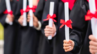 Üniversite kampüsündeki açık hava seremonisi sırasında siyah ve kırmızı mezuniyet kostümleri içinde ellerinde diplomalarla tek sıra bekleyen tanınmamış çok ırklı bir grup.