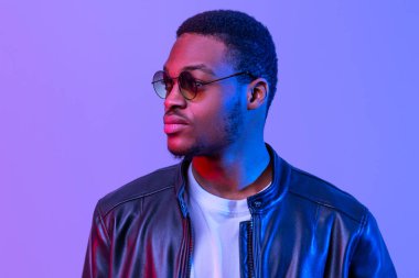 Modaya uygun deri ceketli ve güneş gözlüklü yakışıklı siyah adam portresi Neon Işıklandırma altında poz veren, kendine güvenen Afrikalı Amerikalı adam Mor Stüdyo Arkaplanının üzerinde dikey