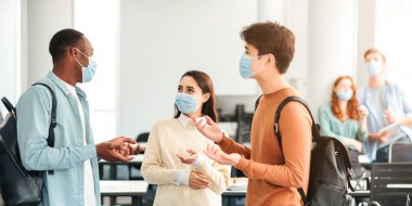 Salgın sırasında güvenli eğitim. Konferans salonunda dikilirken konuşan üç farklı kültürden arkadaşın portresi. Öğrenciler sırt çantaları ve tek kullanımlık yüz maskeleri takıyor. Dersi tartışıyorlar.