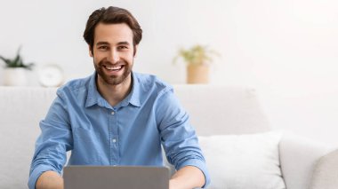 Dizüstü bilgisayardaki mutlu adam internette çalışıyor ve evdeki koltukta oturan kameraya gülümsüyor. Uzaktan kumandalı başarılı adam içeride çalışıyor. Serbest meslek hayatı konsepti