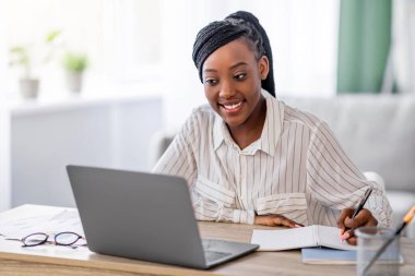Genç siyahi bayan evde online eğitim alıyor, dizüstü bilgisayar kullanıyor ve notlar alıyor, boşluğu kopyalıyor. Gülümseyen Afrikalı Amerikalı kadın çevrimiçi konferansa katılıyor, görevler yazıyor, oturma odası içleri...