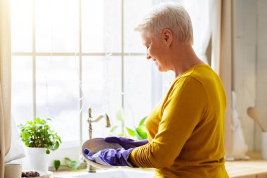 Kıdemli gri saçlı bayan, rahat mutfakta bulaşık yıkıyor, ellerini, arkasını ve fotokopi alanını korumak için lastik eldiven kullanıyor. Evde bulaşık yıkayan çekici bir büyükanne, bezle tabağı temizliyor.