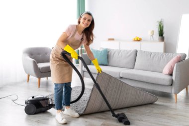 Modern daireyi düzenlemek için elektrikli süpürge kullanan pozitif bir kadın hizmetçi. Ev işlerini yapan, evini düzenli tutan mutlu genç bir kadın. Hijyenik servis kavramı