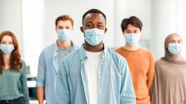 New Reality And Epidemic Concept. Diverse group of international people wearing protective medical masks, standing indoors, posing and looking at camera. Selective focus on african american man