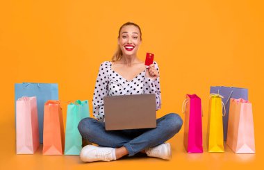 Çevrimiçi Alışveriş Konsepti. Kredi kartı gösteren, dizüstü bilgisayar kullanan, internetten kıyafet ve alışveriş sipariş eden pastel turuncu stüdyoda renkli çantalarla yerde oturan heyecanlı bir kadın.