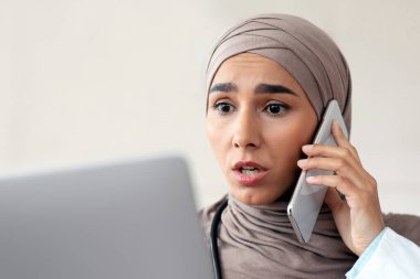 Endişeli kadın doktor tesettürlü genç bayan dizüstü bilgisayara bakıyor, cep telefonuyla konuşuyor, hastayla sohbet ediyor, tavsiye veriyor, klinikte çalışıyor, kopyalama alanında çalışıyor.