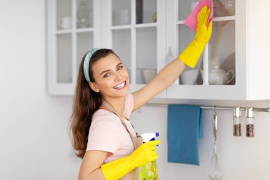 Olumlu kadın temizlikçi modern daireyi düzenliyor, mutfak kabinini deterjan ve bezle siliyor. Ev işleri yapan mutlu genç bir kadın. Profesyonel sağlık hizmeti kavramı