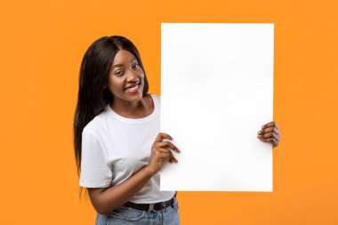 Güzel siyahi bir kadın reklam ya da mesaj için elinde boş bir tahta tutuyor ve turuncu stüdyo arka planında gülümsüyor. Neşeli Afro-Amerikalı kadın dikey boş reklam pankartı gösteriyor.