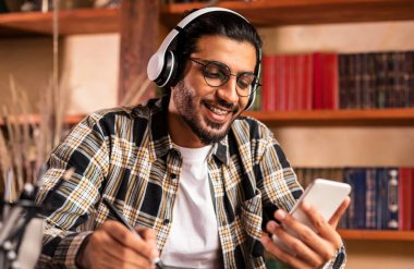 Üniversite kütüphanesinde oturan ve kulaklık takan akıllı telefonlu mutlu Hintli üniversite öğrencisi. Eğitimsel Mobil Uygulama, Yüksek Öğrenim Konsepti