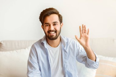 Selamlama Kavramı. Evdeki kanepede otururken el sallayan dost canlısı doğu adamının portresi, olumlu bin yıllık Arap adam gülümsüyor ve kameraya bakıyor, merhaba diyor, boş alan