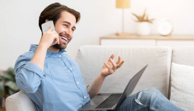 Evdeki koltukta dizüstü bilgisayarla oturup konuşan neşeli adam. Modern İletişim ve Yaşam Tarzı. Mutlu adam cep telefonuyla konuşuyor ve içeride gülüyor.