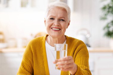 Güzel, yaşlı bir kadın. Kısa gri saçlı, elinde bir bardak su tutan, içi beyaz bir mutfak. Sağlıklı gülümseyen yaşlı kadın. Kaynak suyu, susuzluk konsepti, fotokopi alanı.