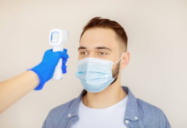 Doktor, klinikte koronavirüs aşısı yapılmadan önce genç adamın sıcaklığını dijital termometreyle ölçüyor. Yüz maskeli bir adam covid-19 aşısı için hastaneyi ziyaret ediyor.