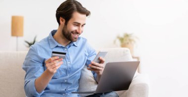 Neşeli adam evde dizüstü bilgisayarla oturup alışveriş yapmak için telefon ve kredi kartı kullanıyor. Mobil Bankacılık, Ödeme Doğrulama, Dükkan Başvurusu Konsepti